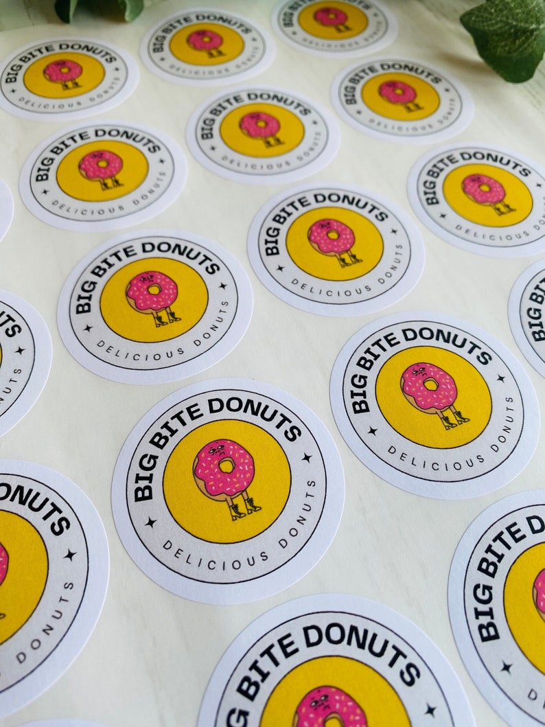 Op de afbeelding: Een verzameling ronde stickers met een gele achtergrond en zwarte tekst die "Big Bite Donuts" en "Delicious Donuts" luidt. Een cartoon-donut met roze glazuur en sprenkels draagt laarzen en heeft een gezicht.