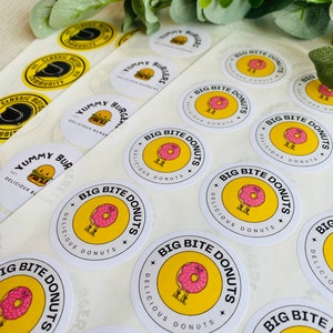 Op de afbeelding: Een vel wit stickerpapier met ronde, gele en witte stickers. De stickers tonen een cartoon-donut met benen en de tekst "Big Bite Donuts" en "Delicious Donuts".