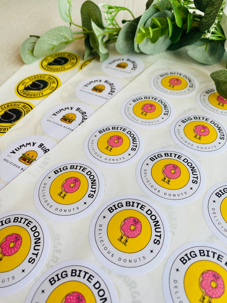 Op de afbeelding: Een vel wit stickerpapier met ronde, gele en witte stickers. De stickers tonen een cartoon-donut met benen en de tekst "Big Bite Donuts" en "Delicious Donuts".