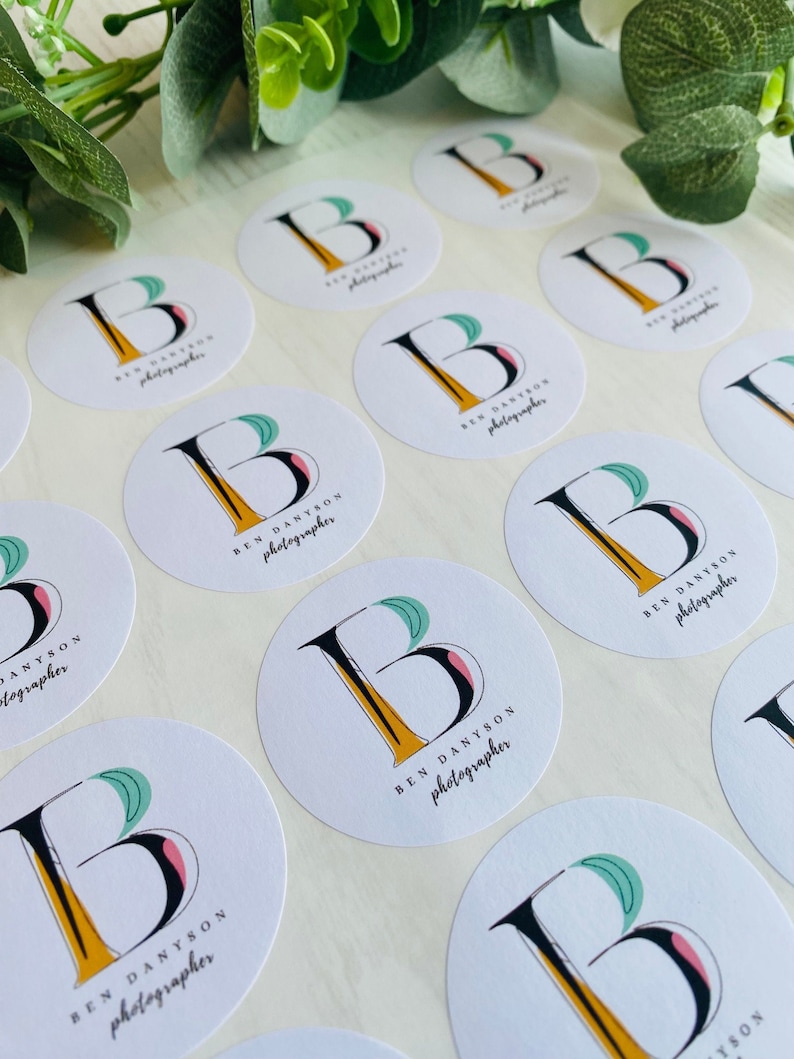 May include: A sheet of white round stickers with a black and yellow stylized letter "B" with a green and pink leaf design. The text "BEN DANYSON photographer" is printed below the letter.