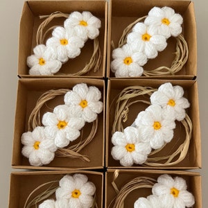 Puede incluir: Seis pequeñas cajas, cada una con tres flores blancas de ganchillo con centros amarillos. Las flores están dispuestas en fila y están anidadas en un material marrón parecido a la paja.