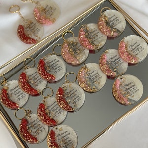 May include: A collection of personalized keychains with gold-colored key rings. The round keychains have a clear resin base with gold flakes and red or pink accents. Each keychain has custom text, including names and dates.