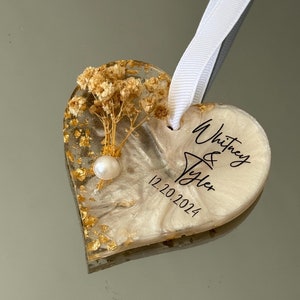 May include: White heart-shaped resin ornament with gold flakes, dried flowers, and a pearl. The ornament is personalized with the names "Whitney & Tyler" and the date "12.20.2024".