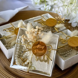 May include: Several white gift boxes with clear lids are arranged on a gold surface. Each box is decorated with a gold circular tag with text, dried flowers, and a gold ribbon. Inside, there are prayer beads and a small cloth.