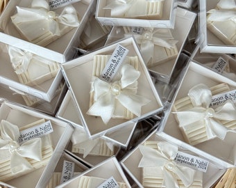 May include: A basket filled with small white boxes, each containing a white soap bar with a white satin ribbon and pearl. The boxes are decorated with a small white card with the text "Manon & Nabil" and the date "septembre 2024".