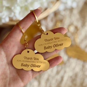 May include: Two gold cloud-shaped keychains with the text "Thank You FOR CELEBRATING WITH US Baby Oliver". The keychains are held up by a person. The background is blurred with white flowers.