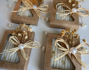 May include: Four small, square, brown boxes with a clear window showing a white bar of soap inside. Each box is decorated with a small bouquet of dried flowers, a pearl, and a brown raffia bow.