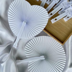 May include: White paper fans, one heart-shaped and one round, are displayed on a white satin surface. The fans are folded in a radial pattern. Additional fans with tags that read "TO OUR BIGGEST FANS Stay Cool" are in a wooden tray.