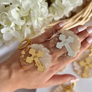 May include: Keyrings featuring gold crosses with the name "Alli" and the year "2025" engraved. The keyrings have a gold ring and are set in clear, glittery resin. White flowers are visible in the background.