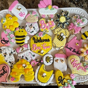 May include: A collection of decorated sugar cookies with a bee and baby shower theme. The cookies are in various shapes, including bees, baby clothes, hearts, and flowers. Colors include yellow, pink, black, and white. Some cookies have text that says "Welcome Ba-bee" and "Sweet Ba-bee".