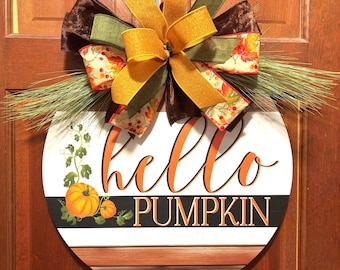 May include: A round wooden door sign featuring the text hello PUMPKIN in orange and black. The sign includes a painted orange pumpkin illustration and a large decorative bow made of brown, green, and orange ribbons with pine needle accents.