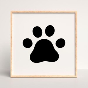 May include: A square wall art piece featuring a black animal paw print centered on a white background, enclosed within a light-colored wooden frame. The design consists of one large central pad and four smaller circular toe pads above it.