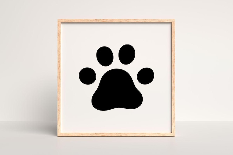 May include: A square wall art piece featuring a black animal paw print centered on a white background, enclosed within a light-colored wooden frame. The design consists of one large central pad and four smaller circular toe pads above it.