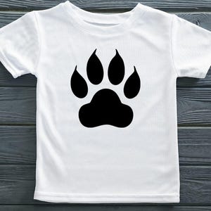 May include: A white short-sleeved t-shirt featuring a large, solid black paw print graphic centered on the chest. The garment has a classic crew neckline and a straight hem, showcasing a simple design with a bold, high-contrast animal motif.