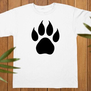 May include: A white short-sleeved t-shirt featuring a large, solid black animal paw print graphic centered on the chest. The design consists of a single large pad at the base with four smaller, rounded toe pads positioned above it in a symmetrical pattern.