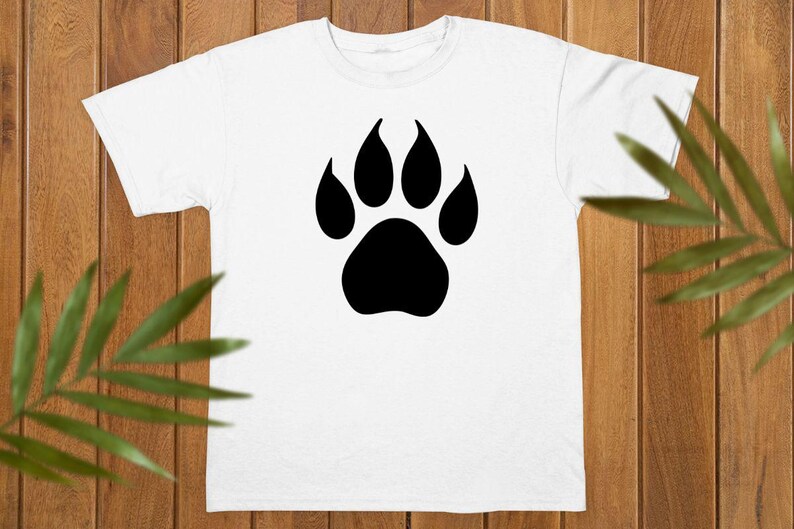 May include: A white short-sleeved t-shirt featuring a large, solid black animal paw print graphic centered on the chest. The design consists of a single large pad at the base with four smaller, rounded toe pads positioned above it in a symmetrical pattern.