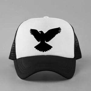 May include: A trucker hat featuring a white front panel with a black silhouette of a bird with wings spread wide. The hat has a solid black brim and black mesh side and back panels, with a button on the top center of the crown for a classic look.