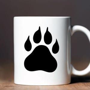 May include: A white ceramic mug featuring a large, solid black animal paw print graphic centered on the front. The mug has a smooth, cylindrical body and a curved handle on the right side, providing a clean and simple design for daily beverage use.