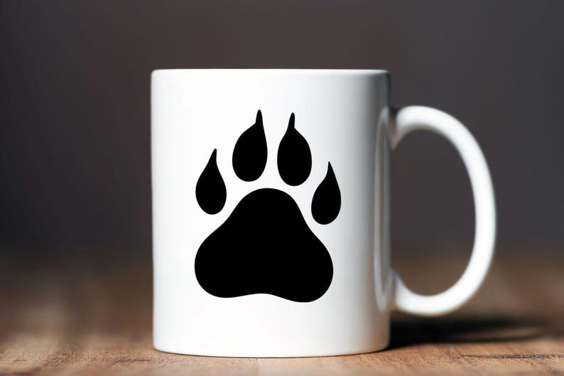 May include: A white ceramic mug featuring a large, solid black animal paw print graphic centered on the front. The mug has a smooth, cylindrical body and a curved handle on the right side, providing a clean and simple design for daily beverage use.
