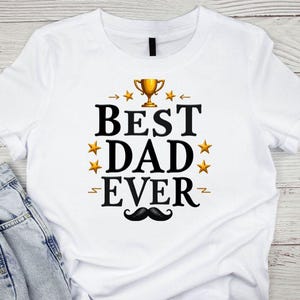 May include: White t-shirt with the text "BEST DAD EVER" in black, gold, and yellow. The design includes a gold trophy, gold stars, and a black mustache. The shirt is paired with blue jeans.