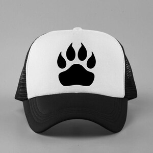 May include: A trucker hat featuring a white front panel with a black paw print graphic, a solid black brim, and black mesh side and back panels. The cap has a structured front and an adjustable snapback closure visible on the rear side.