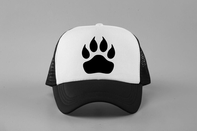 May include: A trucker hat featuring a white front panel with a black paw print graphic, a solid black brim, and black mesh side and back panels. The cap has a structured front and an adjustable snapback closure visible on the rear side.