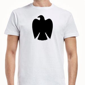 May include: A white short-sleeved t-shirt featuring a solid black silhouette of an eagle with its wings tucked in, centered on the chest. The garment has a crew neckline and a standard fit, providing a clean contrast between the dark graphic and fabric.