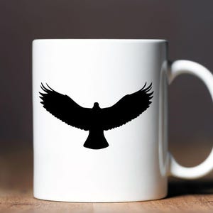 May include: A white ceramic mug featuring a black silhouette of a bird with its wings fully spread in flight. The bird is centered on the front of the mug, which has a smooth, cylindrical body and a curved handle on the right side.