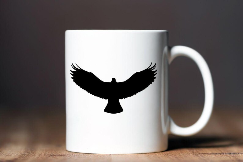 May include: A white ceramic mug featuring a black silhouette of a bird with its wings fully spread in flight. The bird is centered on the front of the mug, which has a smooth, cylindrical body and a curved handle on the right side.