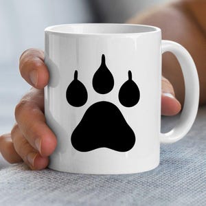 May include: A white ceramic mug featuring a large, solid black paw print graphic centered on the front. The mug has a smooth, cylindrical body with a curved handle on the right side, providing a clean and simple design for holding hot beverages.