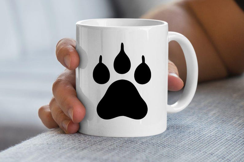May include: A white ceramic mug featuring a large, solid black paw print graphic centered on the front. The mug has a smooth, cylindrical body with a curved handle on the right side, providing a clean and simple design for holding hot beverages.