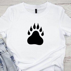 May include: A white short-sleeved t-shirt featuring a large, solid black bear paw print graphic centered on the chest. The shirt has a classic crew neckline and is paired with light blue denim jeans visible at the bottom left corner.