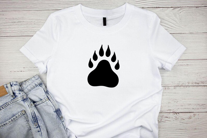 May include: A white short-sleeved t-shirt featuring a large, solid black bear paw print graphic centered on the chest. The shirt has a classic crew neckline and is paired with light blue denim jeans visible at the bottom left corner.