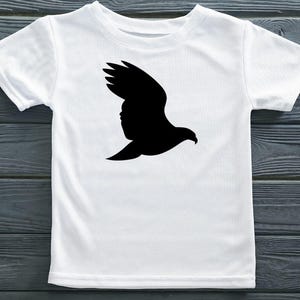 May include: A white short-sleeved t-shirt featuring a solid black silhouette of a bird in flight centered on the chest. The garment has a crew neckline and a standard fit, showcasing a clean contrast between the dark graphic and the light fabric.