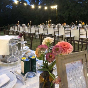 May include: A table setting for an outdoor wedding reception with white linens,  a brown vase with pink and yellow flowers, two green olive oil bottles, and a wooden frame with a menu titled "Menu".
