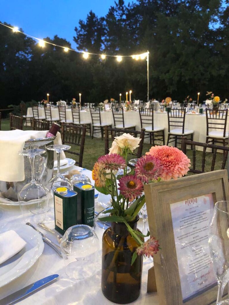 May include: A table setting for an outdoor wedding reception with white linens,  a brown vase with pink and yellow flowers, two green olive oil bottles, and a wooden frame with a menu titled "Menu".