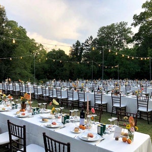 May include: An outdoor wedding reception with white linens and chairs set up on a grassy lawn. The tables are decorated with flowers and candles. String lights are strung across the lawn, creating a romantic ambiance.