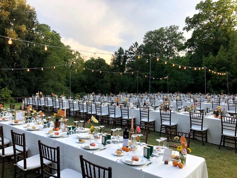 May include: An outdoor wedding reception with white linens and chairs set up on a grassy lawn. The tables are decorated with flowers and candles. String lights are strung across the lawn, creating a romantic ambiance.