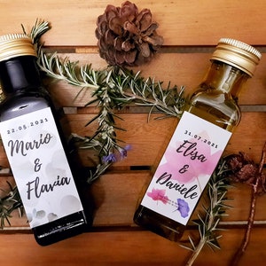 May include: Two small glass bottles with labels featuring watercolor floral designs and the names Mario & Flavia and Elisa & Daniele. The bottles are on a wooden surface with sprigs of rosemary and pine cones.