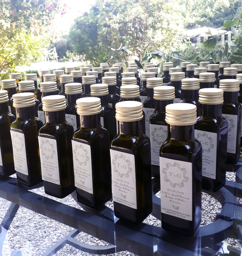 May include: A collection of small glass bottles with olive oil, each with a personalized label featuring a monogram and the text "T.G. 2023 Villa Sant'Anna Extra Virgin Olive Oil".