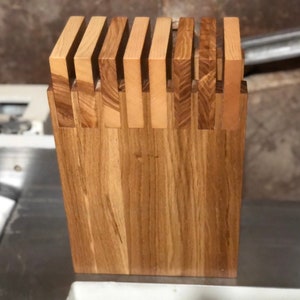 May include: A wooden knife block with a natural wood grain finish. The block has vertical slots for knife storage, and a dovetail joint construction. The wood has a warm, light brown colour, and is likely made from oak.