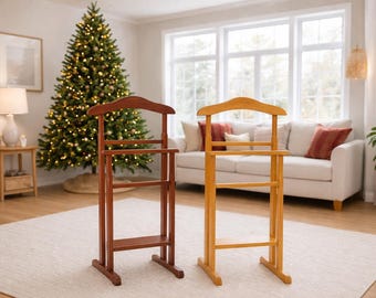 May include: Two wooden valet stands, one in a rich brown and the other in a light honey color, stand side-by-side on a neutral rug. Each has a curved top, a bar for hanging clothes, and a lower shelf. The background includes a Christmas tree.