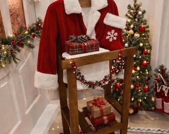 May include: A red Santa Claus suit with white faux fur trim is displayed on a wooden stand, alongside a red Christmas stocking with a snowflake design. Several wrapped gifts and a small Christmas tree are also present, creating a festive holiday scene.