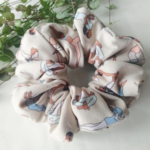 May include: A beige scrunchie with a repeating pattern of women in various yoga poses. The women are illustrated in shades of brown, pink, and blue.