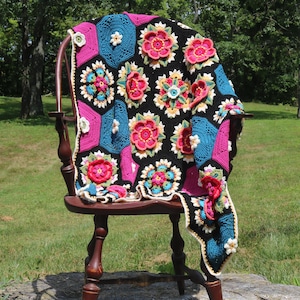 May include: A colorful crocheted throw blanket with a floral pattern in shades of pink, blue, yellow, and green. The blanket is draped over a wooden chair.
