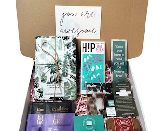 May include: A gift box filled with various items, including a book wrapped in green and white patterned paper, a chocolate bar with the words "HIP! Oat Milk Chocolate" and a goat graphic, a bookmark with the text "Once you learn to read, you will be forever free", two tea bags, a small card with the text "Happily Ever After Book Box", a biscuit, and a card with the text "Thank you. The box has raised money for..."
