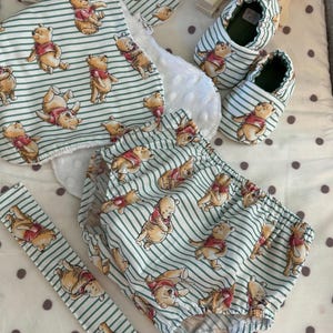 May include: A Winnie the Pooh baby outfit set with a green and white striped pattern. The set includes a bib, a pair of booties, a diaper cover, and a teether. The outfit is perfect for a baby shower gift.