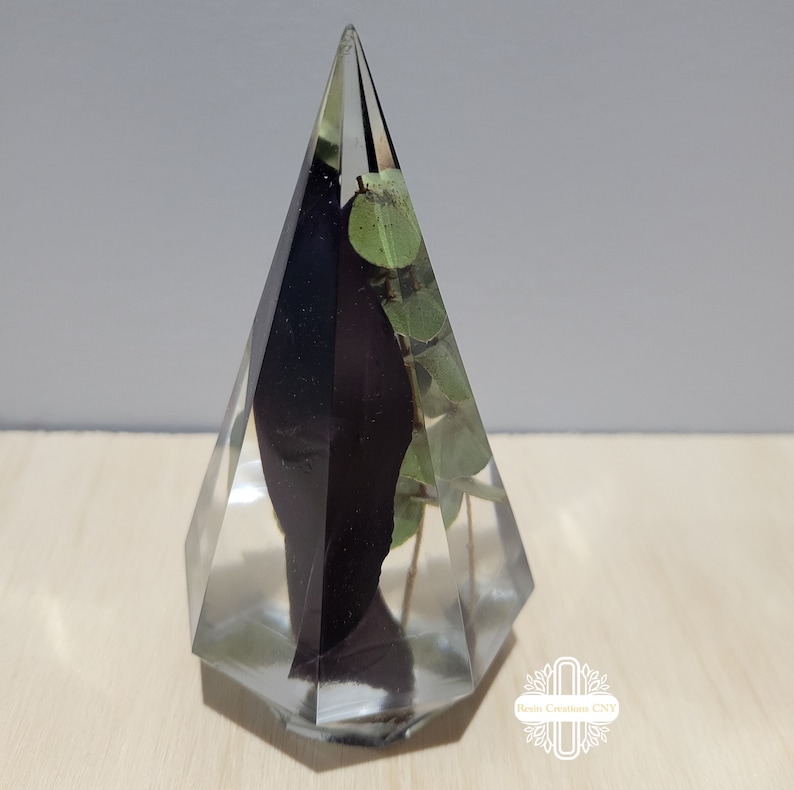 May include: A clear glass pyramid with a black flower and green leaves preserved inside. The pyramid is on a light wood surface.
