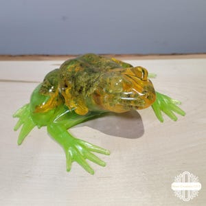 Może przedstawiać: P&oacute;łprzezroczysta figurka żaby z żywicy o żywej, limonkowo-zielonej podstawie i teksturowanym grzbiecie w kolorach mchu i bursztynu. Rzeźba posiada szczeg&oacute;łowe błony pławne oraz gładkie, błyszczące wykończenie z wyraźnie zaznaczonym okiem.