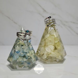 May include: Two clear resin ring holders shaped like pyramids. The holders are filled with dried flowers. One holder has white flowers and the other has blue and white flowers. The holders are on a white surface.